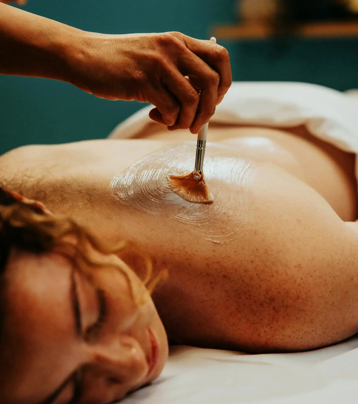 A therapist brushing a treatment mask onto a guest's back in a teal treatment room